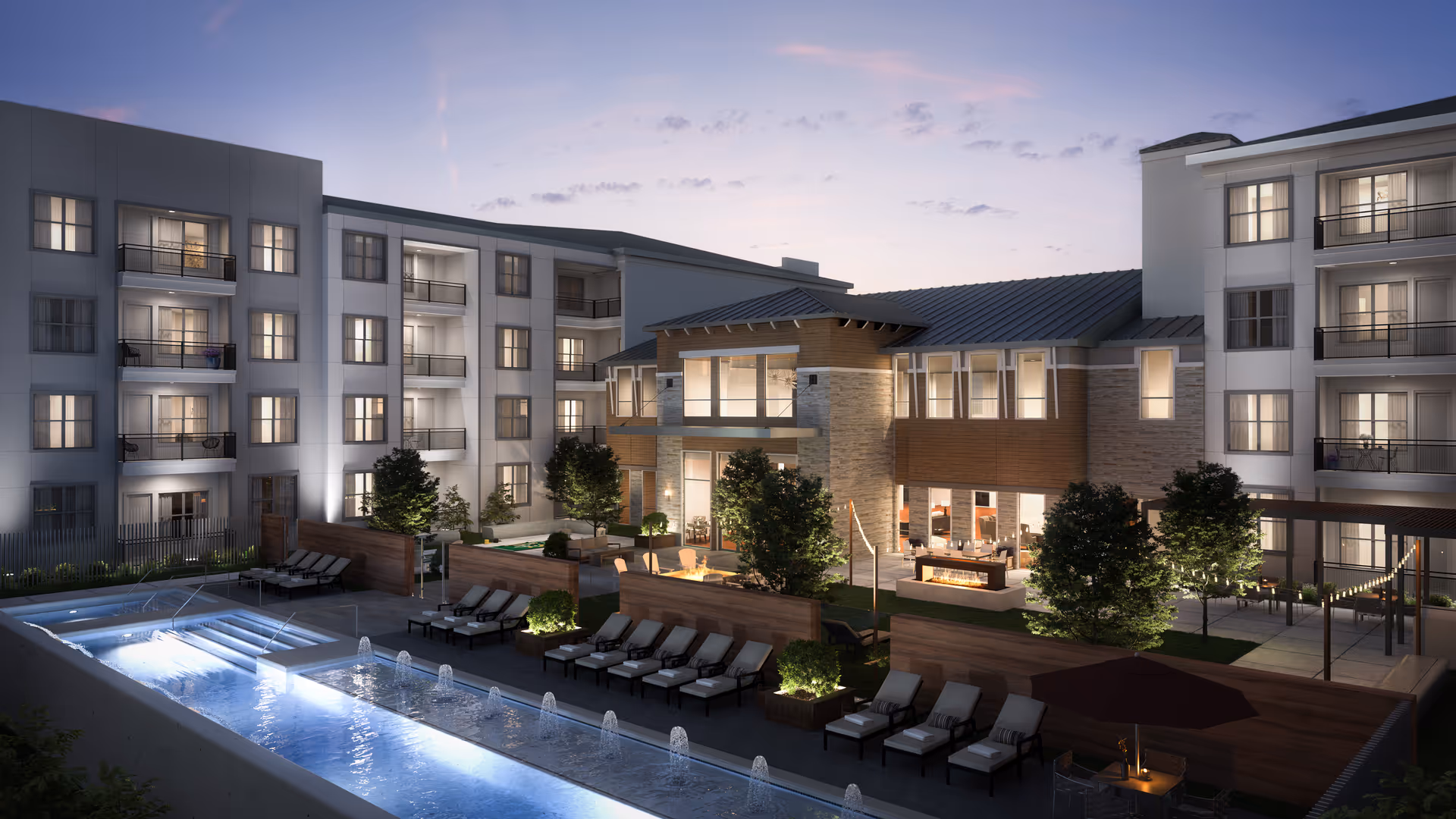 Dusk view of an outdoor courtyard with a long illuminated pool, lounge chairs, fountains and a multi-story residential building with balconies.
