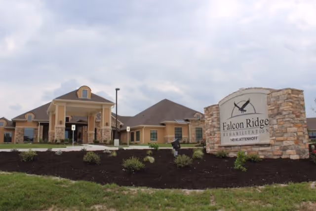 Exterior view of Falcon Ridge Rehabilitation facility showing a single-story building with a covered entrance, stone and beige siding, and a landscaped area with shrubs and grass in front. A large stone sign with the facility name and address is visible on the right side.