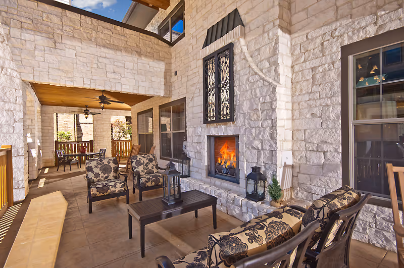 Outdoor covered patio area with stone walls and a lit fireplace. The space includes cushioned chairs and a coffee table with lanterns, a ceiling fan, and a dining table with chairs in the background. The area is well-lit with natural sunlight.