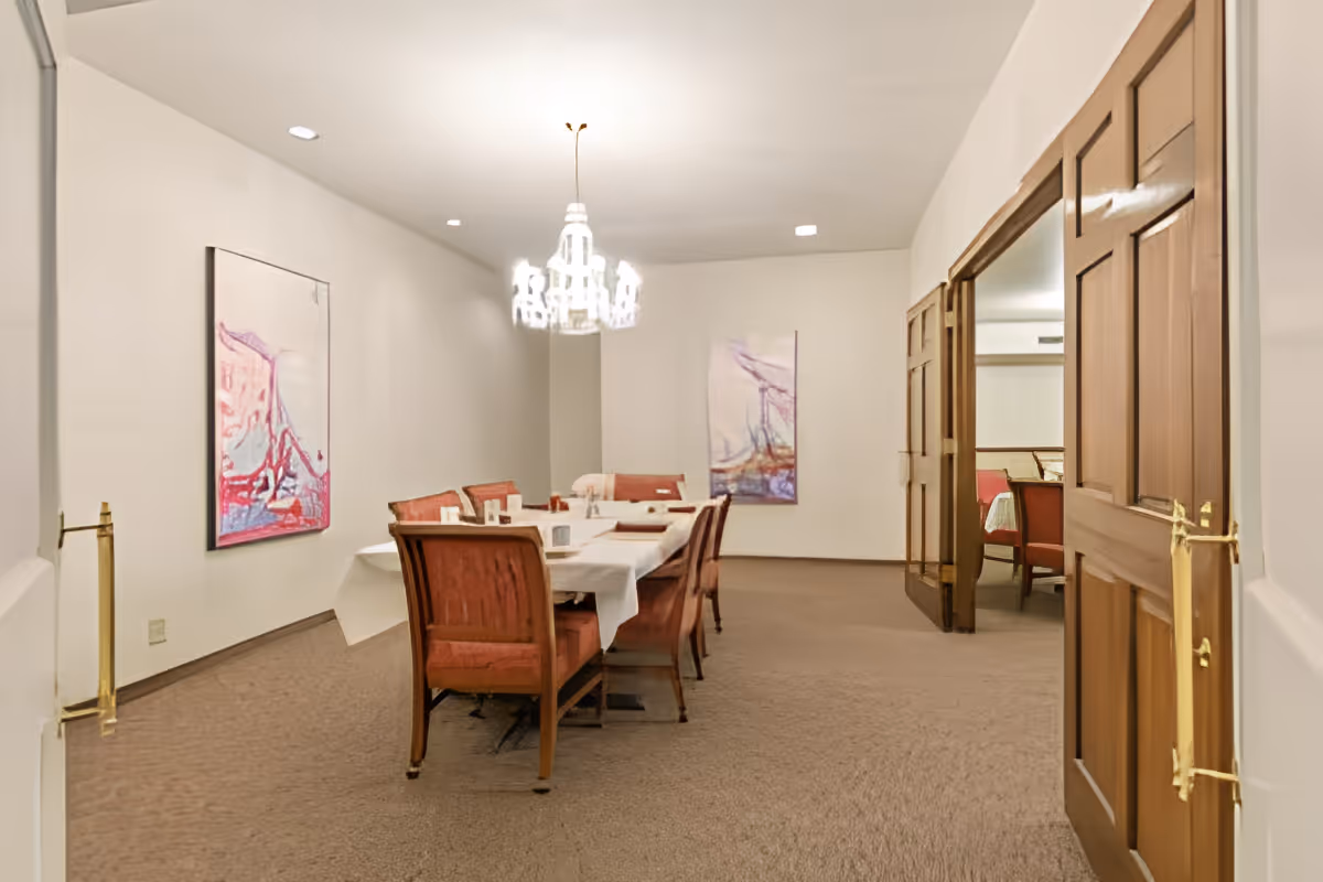 A dining room with a long table covered with a white tablecloth and surrounded by wooden chairs with red cushions. Two abstract paintings hang on the walls, and a chandelier is centered above the table. The room has beige carpet and wooden double doors that lead to another room with similar decor.