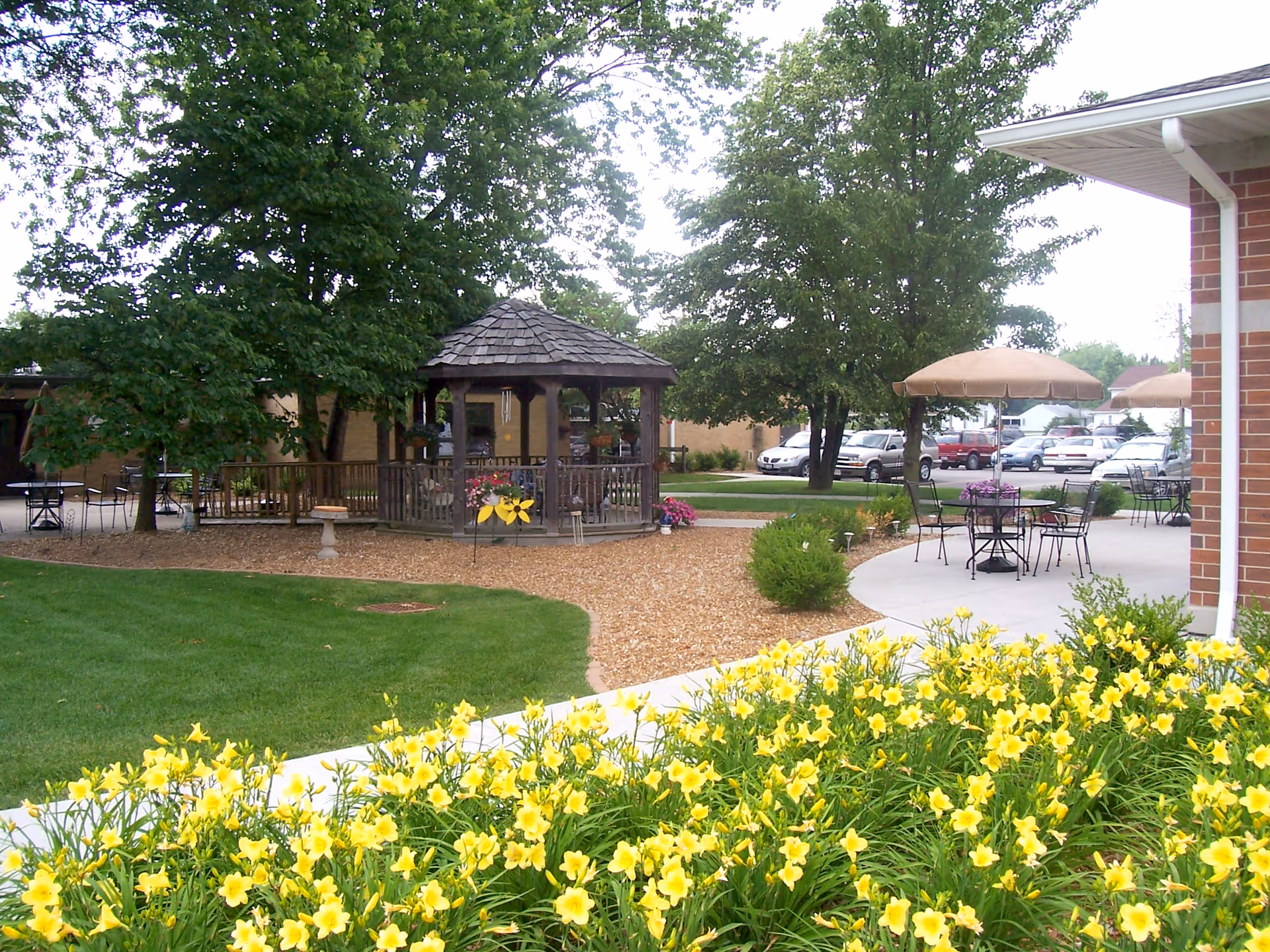 Outdoor garden area with a wooden gazebo surrounded by trees and flowers. There are patio tables with umbrellas and chairs on a concrete surface near a brick building. Yellow flowers are blooming in the foreground, and cars are parked in the background.