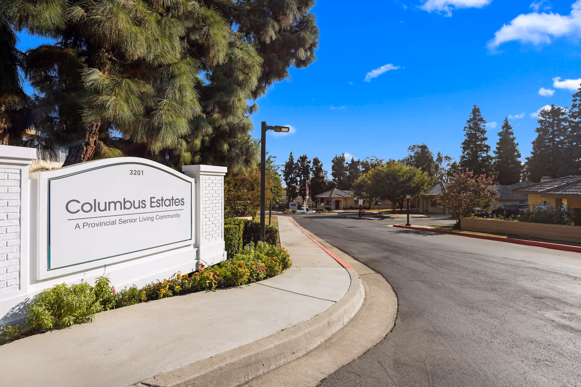Entrance sign for Columbus Estates, a Provincial Senior Living Community, situated next to a curved driveway with trees and buildings in the background under a clear blue sky.