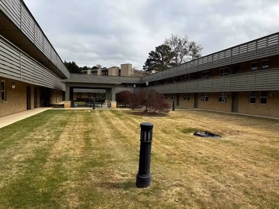 Outdoor courtyard between two wings of a senior living facility, with a central lamp post, a small tree, and a seating area in the distance.