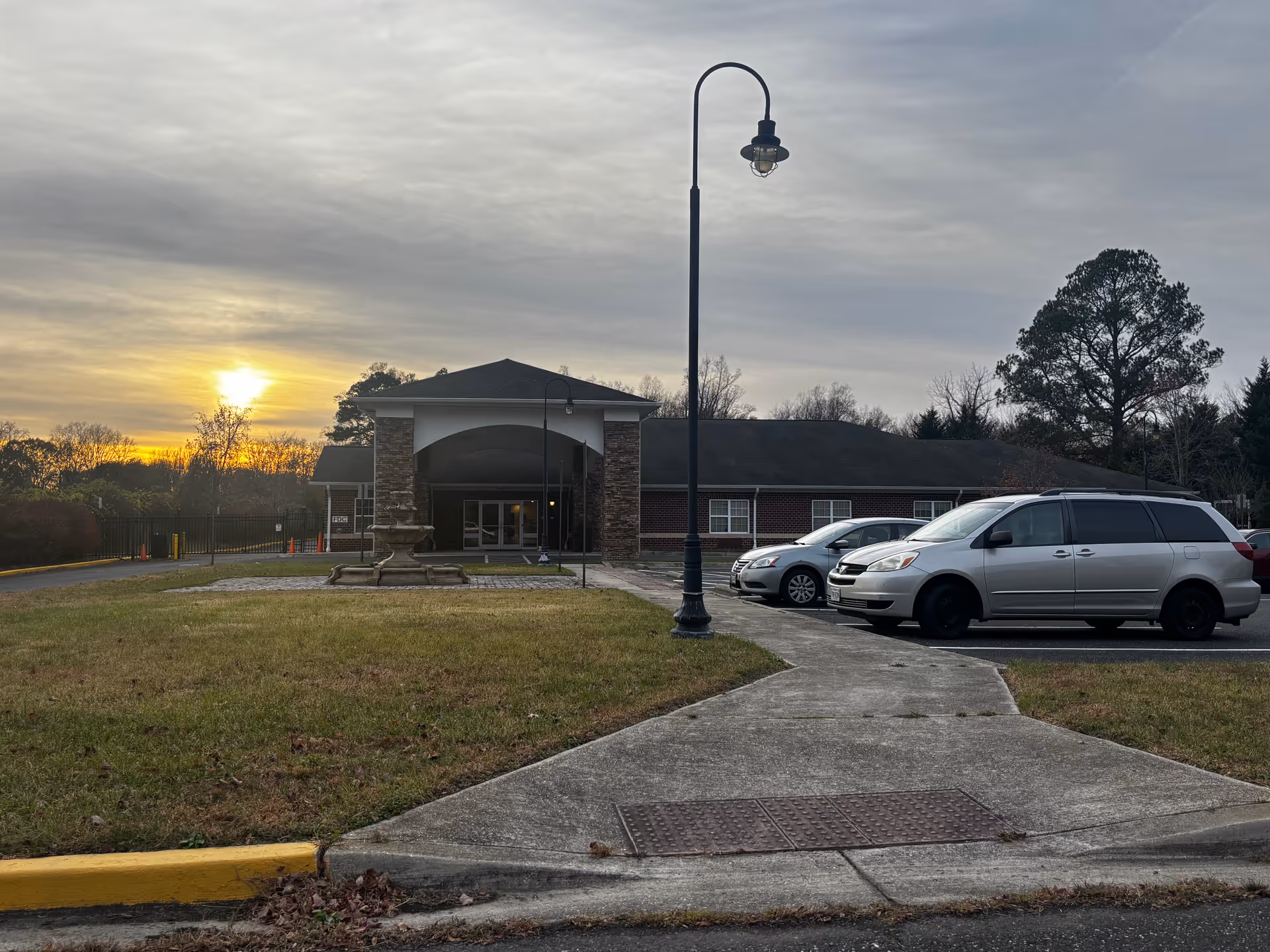 Exterior view of The W Assisted Living facility at sunset, showing a single-story brick building with a covered entrance supported by stone pillars. There is a concrete walkway leading to the entrance, a street lamp, a small fountain in front of the building, and several parked cars in the parking lot. Trees and a cloudy sky are visible in the background.