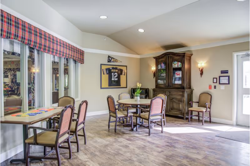 A well-lit common area with a round table and chairs, a cabinet with glass doors, and a framed sports jersey on the wall. There are windows with plaid valances and a door letting in natural light.