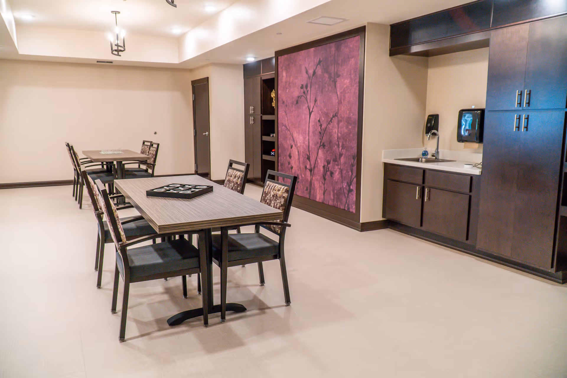 A clean and modern dining area with two rectangular tables, each surrounded by four chairs with patterned backs. The room features light-colored flooring, beige walls, and dark wood cabinetry along one wall with a small sink and soap dispenser. A large decorative panel with a purple floral design is mounted on the wall. The space is well-lit with ceiling lights.