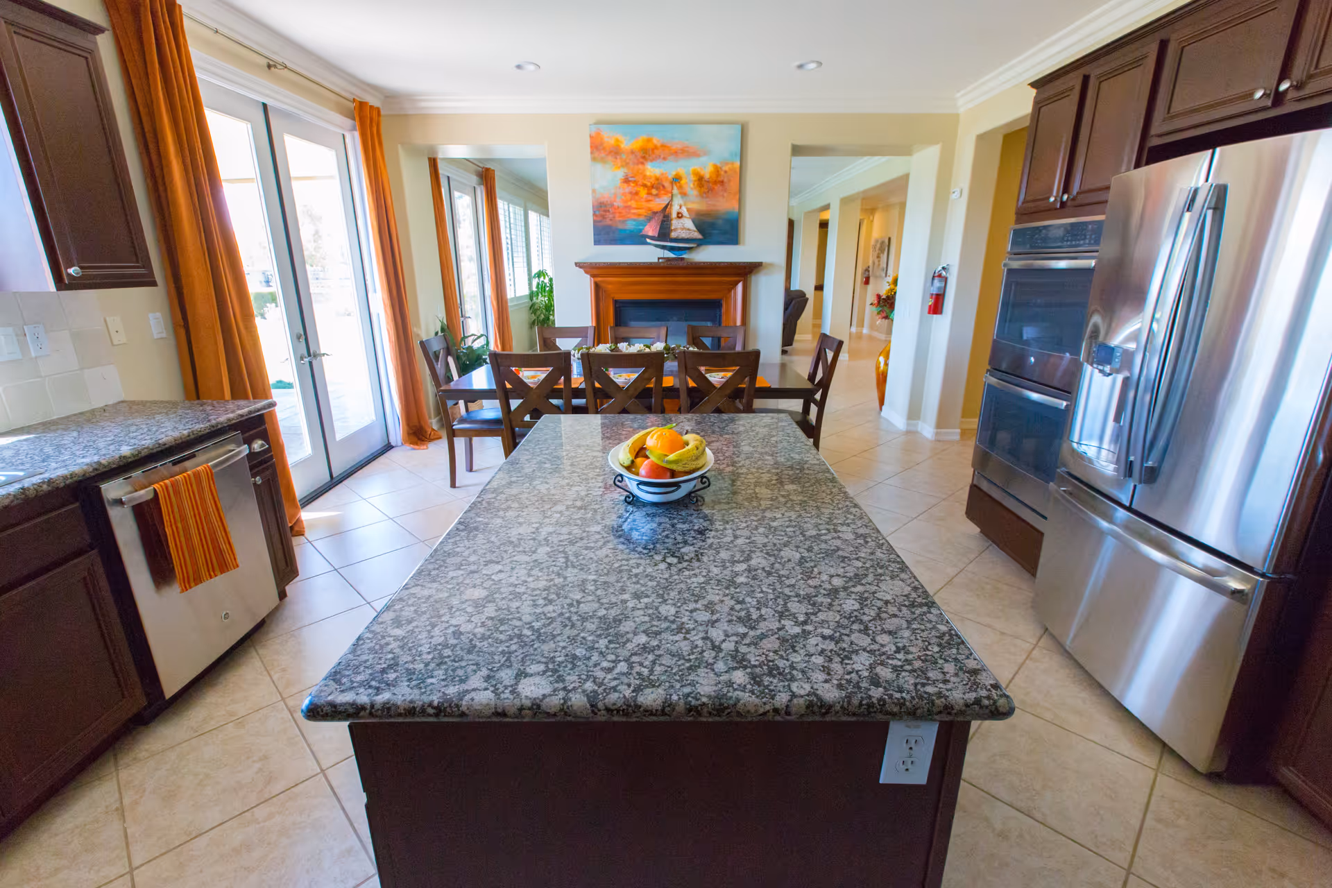 A bright kitchen and dining area with a large granite island countertop in the foreground holding a bowl of fruit. The kitchen features dark wood cabinets, stainless steel appliances including a refrigerator and double oven, and a dishwasher with an orange striped towel. The dining area has a wooden table with six chairs, orange curtains on glass doors leading outside, and a fireplace with a colorful painting above it.