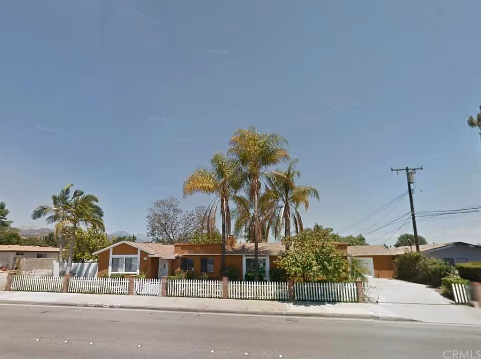 Single-story residential building with a white picket fence in front, several palm trees and other greenery around the property, under a clear blue sky.