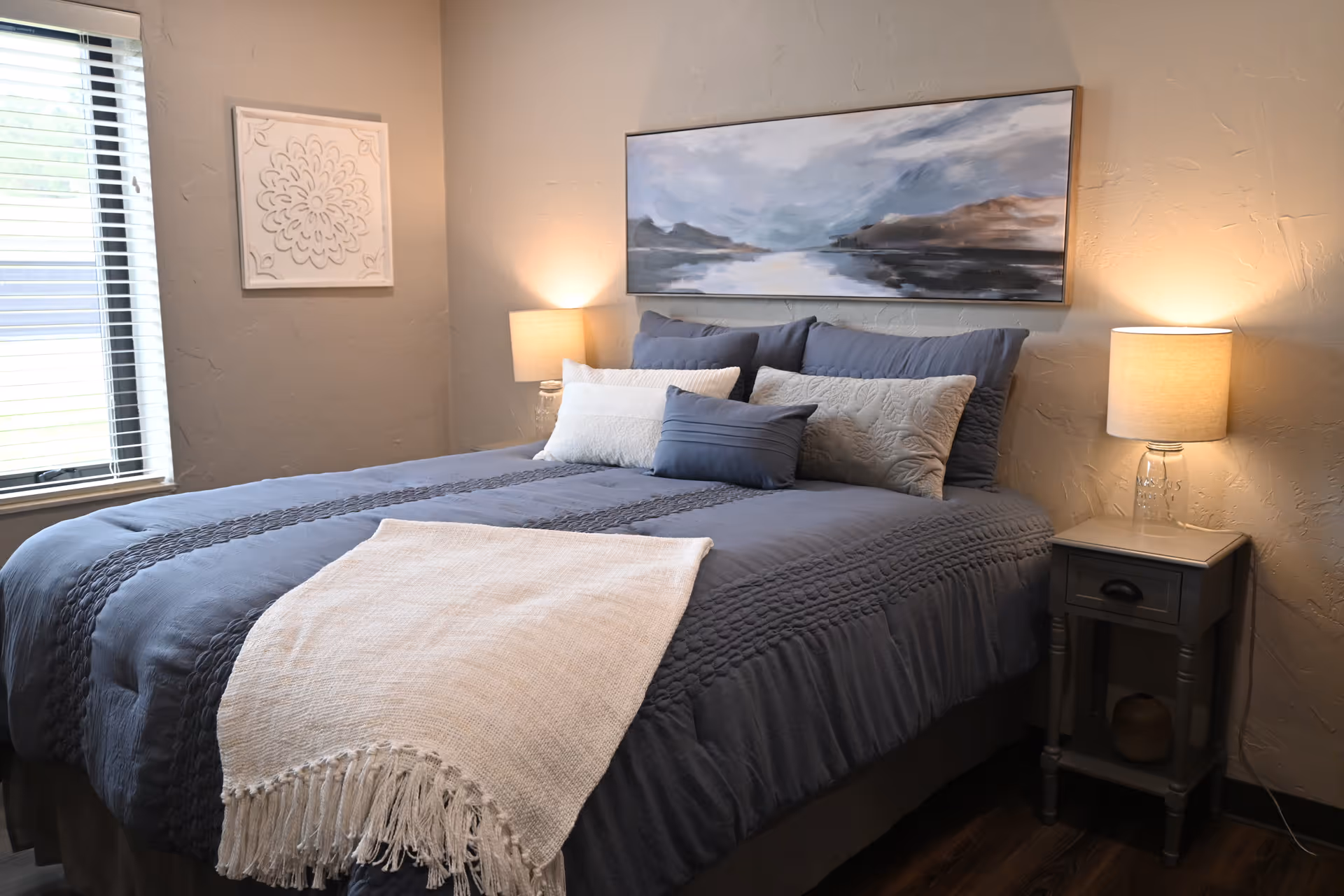 A cozy bedroom with a neatly made bed featuring blue and white bedding and multiple pillows. There are two bedside tables with lamps on each side of the bed. A large abstract painting hangs above the bed, and a decorative wall piece is on the adjacent wall near a window with blinds.
