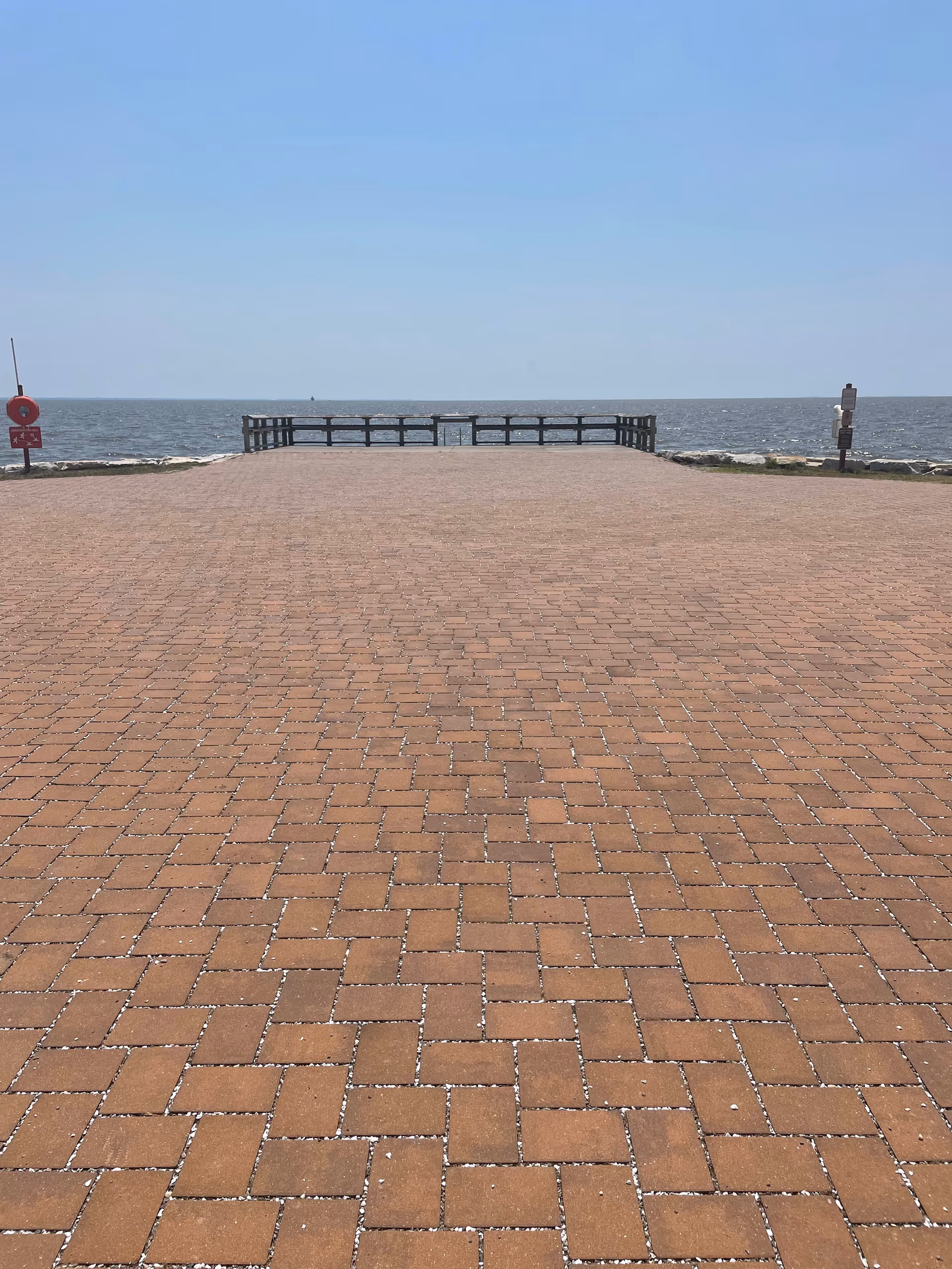 A paved brick walkway leading to a wooden railing overlooking a large body of water under a clear blue sky.