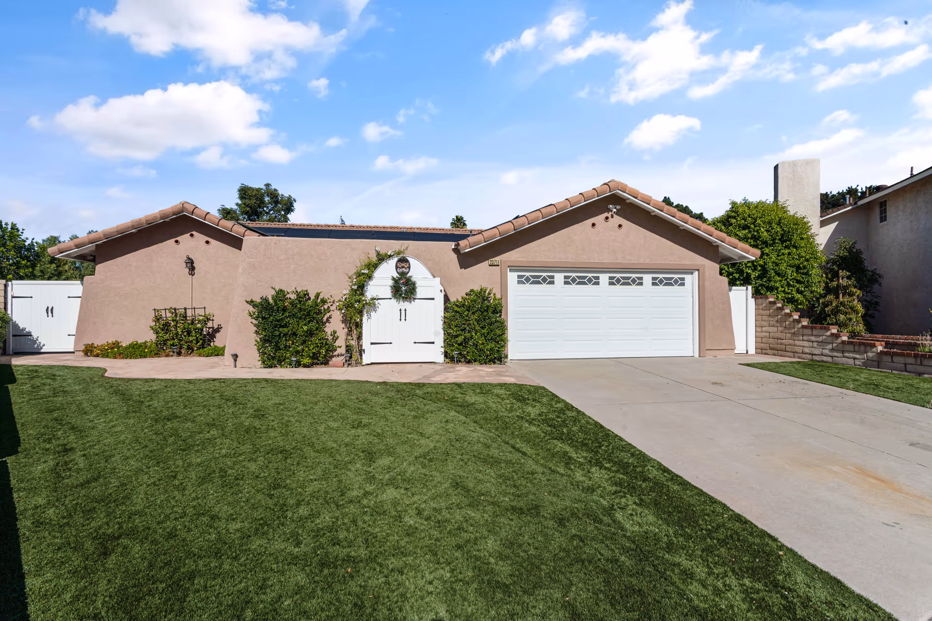 Front exterior view of a single-story house with a beige stucco finish and a tiled roof. The house features a white double garage door on the right and a white arched gate decorated with a wreath in the center. There are green bushes on either side of the gate and a well-maintained green lawn in front. The sky is blue with scattered clouds.