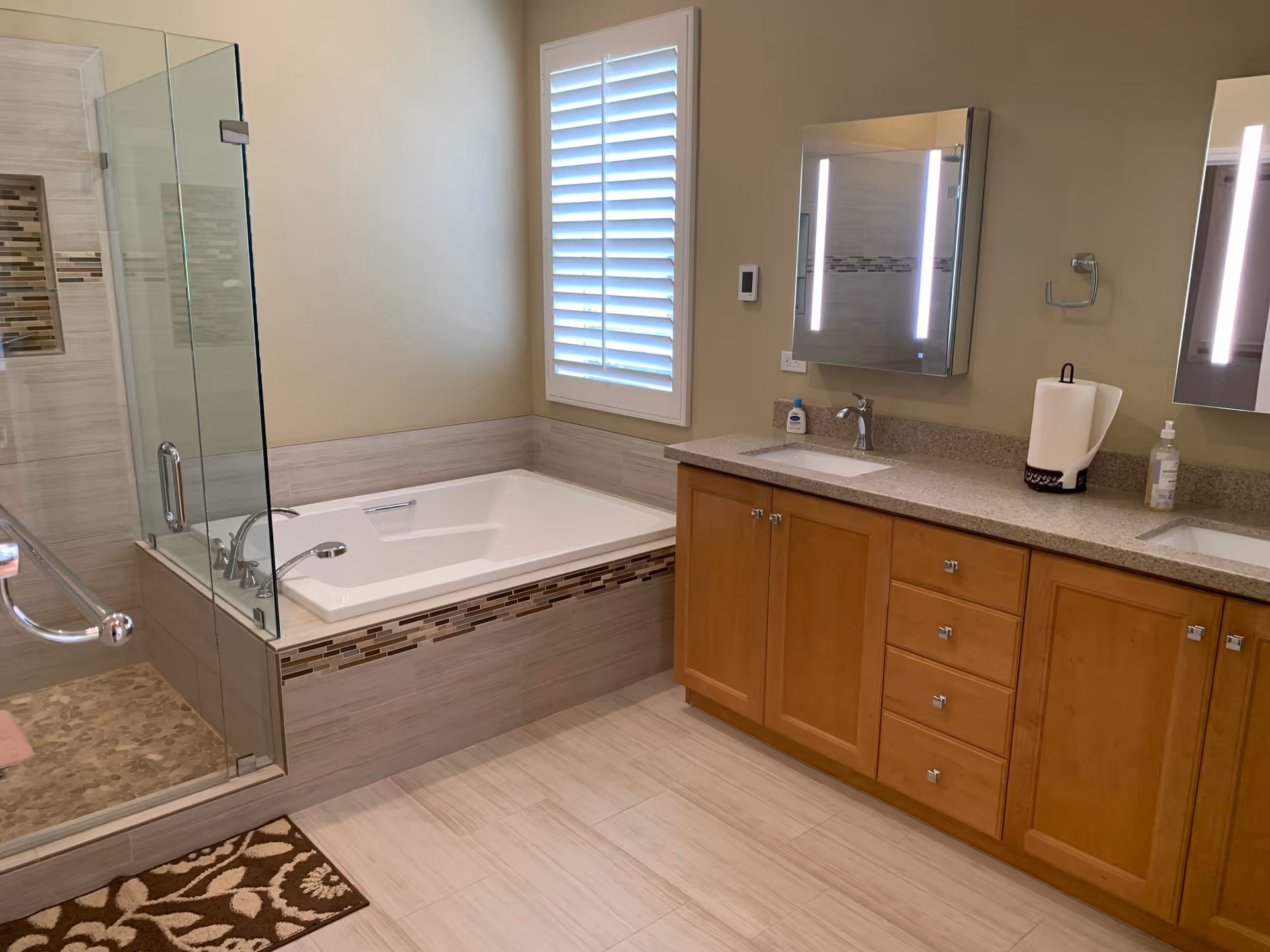 A modern bathroom featuring a large bathtub with tiled surround and a glass-enclosed shower area. There is a window with white plantation shutters above the bathtub. A double sink vanity with wooden cabinets and illuminated mirrors is on the right side. The floor is tiled and there is a patterned brown and beige rug near the shower entrance.