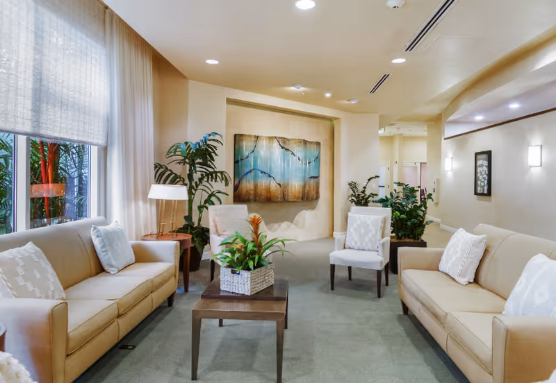 A well-lit living room area with two beige sofas, two cushioned chairs, a wooden coffee table with a potted plant, large windows with sheer curtains, indoor plants, and abstract wall art.