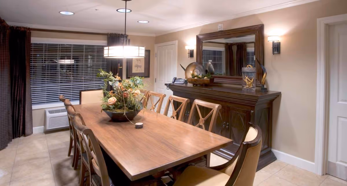 A dining room with a long wooden table surrounded by eight chairs. A large floral centerpiece is on the table. The room has beige walls, a large mirror above a dark wooden sideboard, and a window with blinds and dark curtains. Ceiling lights and wall sconces illuminate the space.