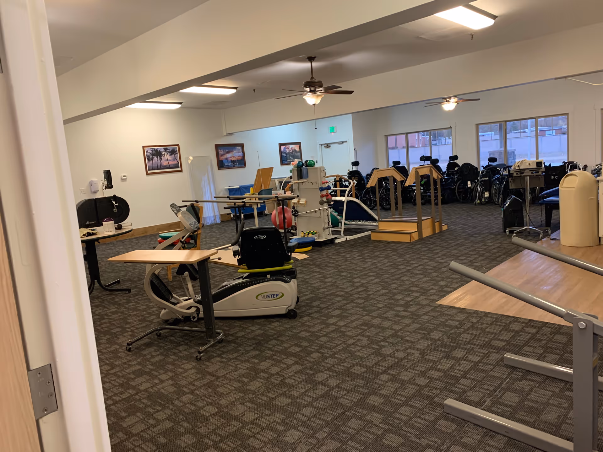 A spacious rehabilitation room with exercise equipment including a recumbent bike, parallel bars for walking practice, and various other physical therapy tools. The room has carpeted flooring, ceiling fans, and several windows letting in natural light. Wheelchairs are lined up against the far wall.