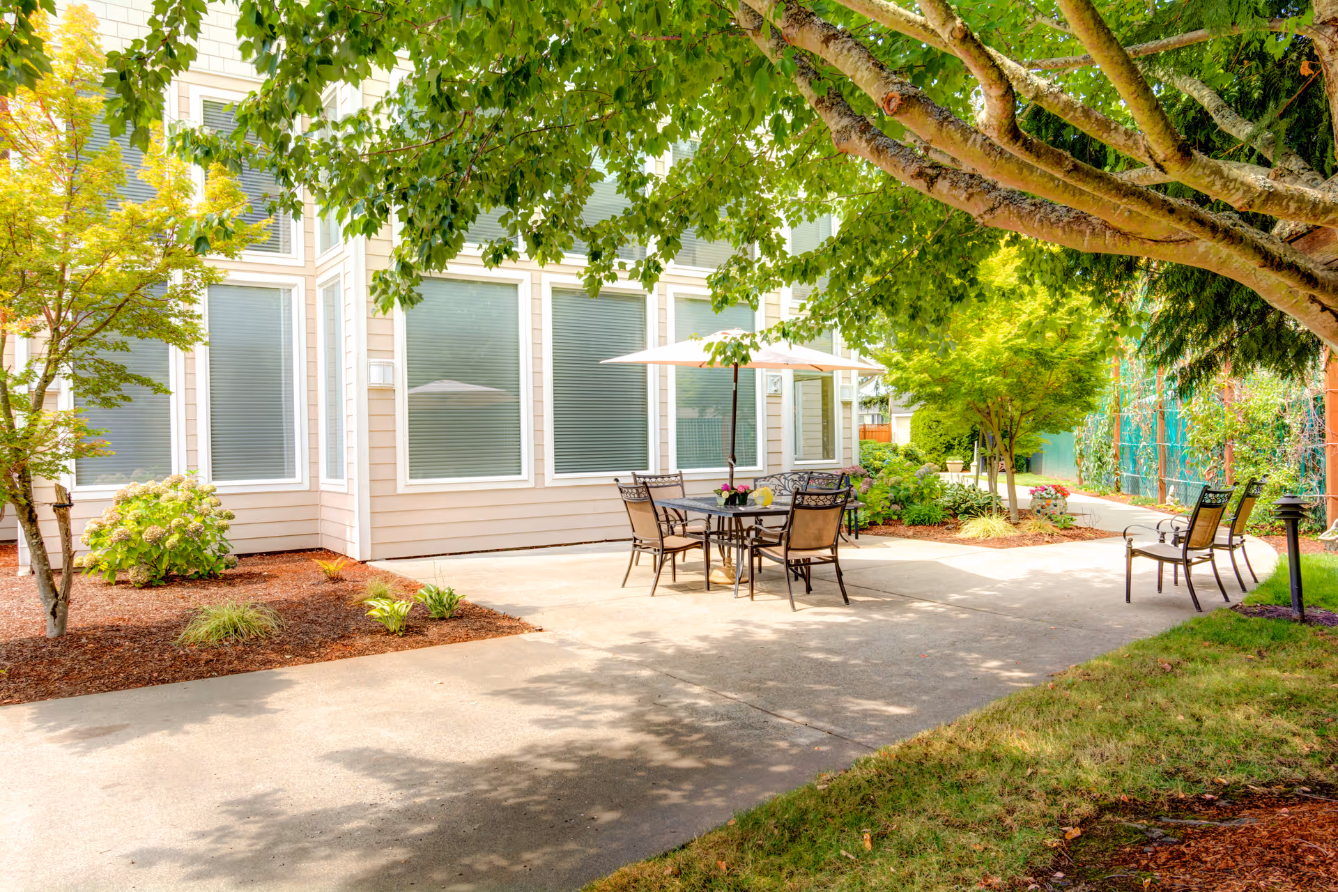 Outdoor patio area at Cedar Village by Cogir featuring a table with an umbrella and chairs surrounded by trees and greenery, adjacent to a building with large windows.