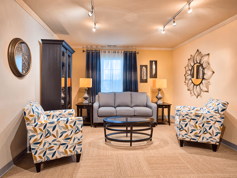 A cozy living room with a gray sofa centered against a wall with a window covered by dark blue curtains. On either side of the sofa are dark wooden side tables with matching lamps. Two patterned armchairs with blue and yellow leaf designs face the sofa. A round glass coffee table with a black metal frame sits in the middle on a beige carpet. The walls are decorated with a round mirror with a sunburst frame and two framed leaf artworks. A dark wooden cabinet stands to the left of the sofa.