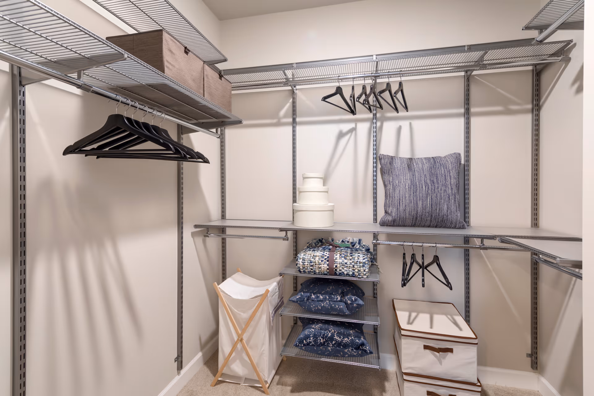 A spacious walk-in closet with metal shelving and hanging rods. The closet contains several black hangers, a few decorative pillows, a folded blanket, a laundry hamper with a wooden frame, and a fabric storage box. The walls are painted light beige and the floor is carpeted.