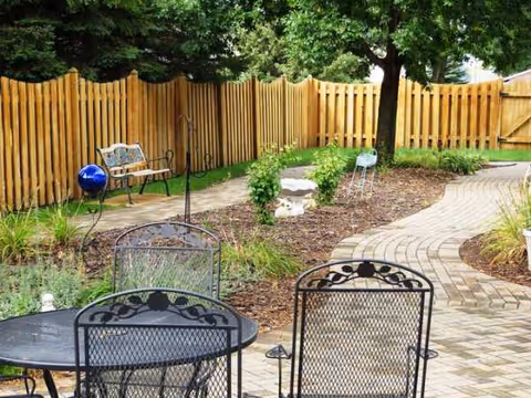 Outdoor patio features metal chairs, a brick path, garden beds, and wooden fencing.