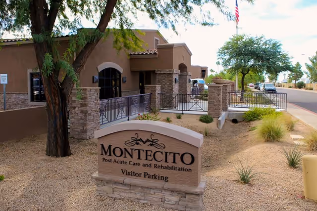 Exterior view of Montecito Post Acute Care and Rehabilitation facility showing a stone and stucco building with a sign in front indicating visitor parking. There are trees, shrubs, and a paved road with parked cars in the background.