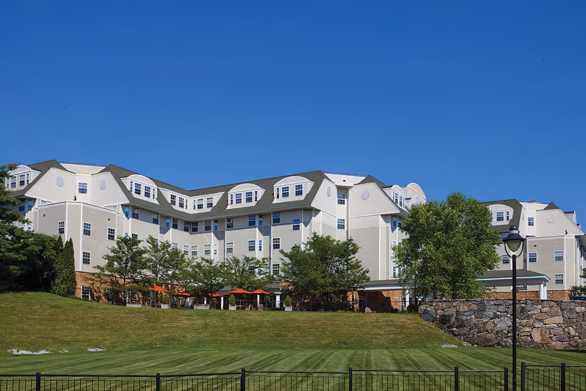 Multi-story senior living building with a manicured lawn, trees, outdoor seating and a clear blue sky.