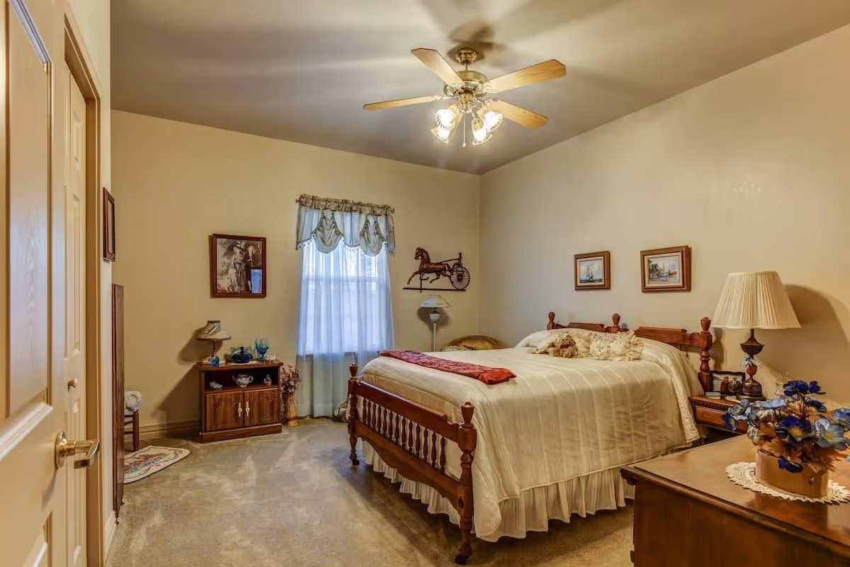 Traditional bedroom with a wooden bed, bedside tables, a ceiling fan, and a curtained window.