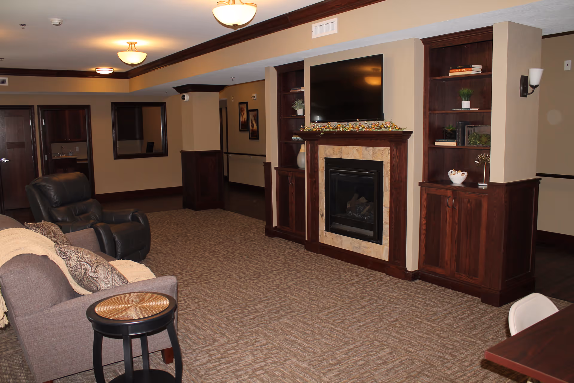 A cozy living room area in a senior living facility featuring a fireplace with a mounted flat-screen TV above it, flanked by built-in wooden shelves with decorative items. There are two comfortable chairs, a small round side table, and warm lighting from ceiling fixtures. The walls are painted beige with dark wood trim, and the floor is carpeted.