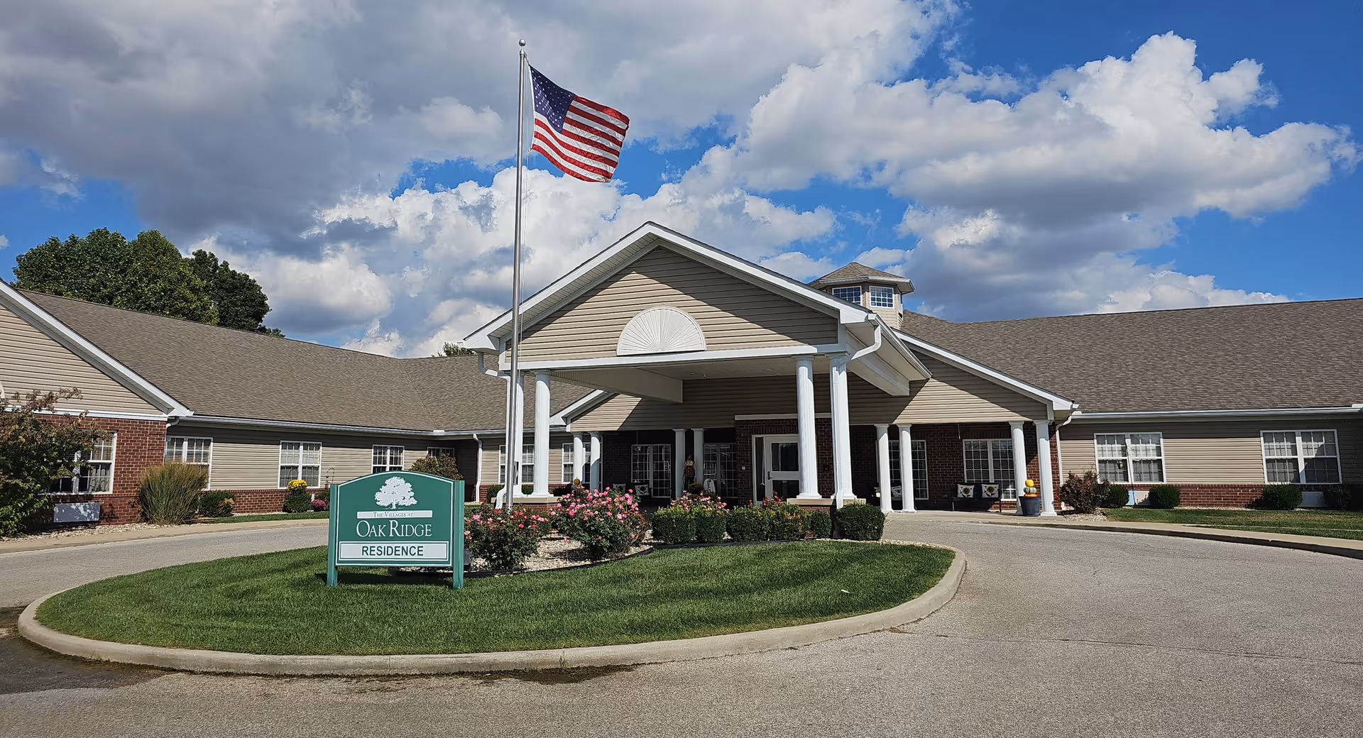 Exterior front entrance of The Villages at Oak Ridge residence with a covered portico, American flag, circular driveway, and landscaping.