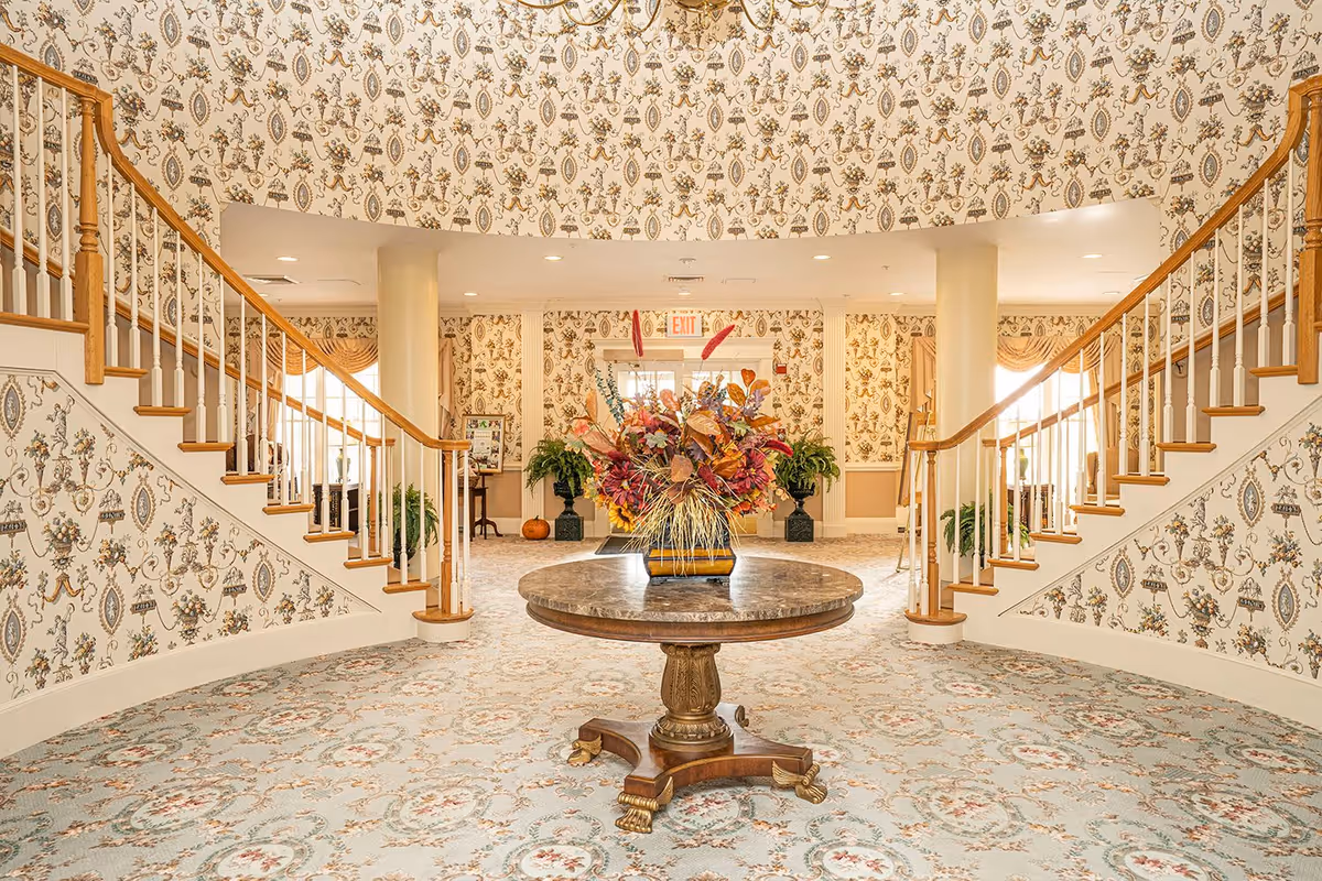 A grand interior entrance hall with a round wooden table in the center holding a large floral arrangement. The room features a patterned carpet and wallpaper with a vintage design. Two symmetrical staircases with wooden handrails and white balusters curve upward on either side of the room. There are columns and potted plants near the back wall, with windows allowing natural light to enter.