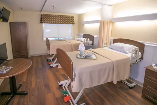 A rehabilitation center room with two hospital beds, each with beige bedspreads and white pillows. One bed has a tray with a napkin, plate, and cup. The room has wood flooring, a small round table with magazines, a wooden cabinet, a wall-mounted TV, and a window with a patterned valance. A privacy curtain is partially drawn between the beds.