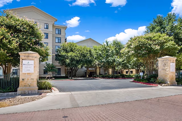 Driveway entrance and landscaped front of a multi-story building with stone pillars, trees, and a blue sky.
