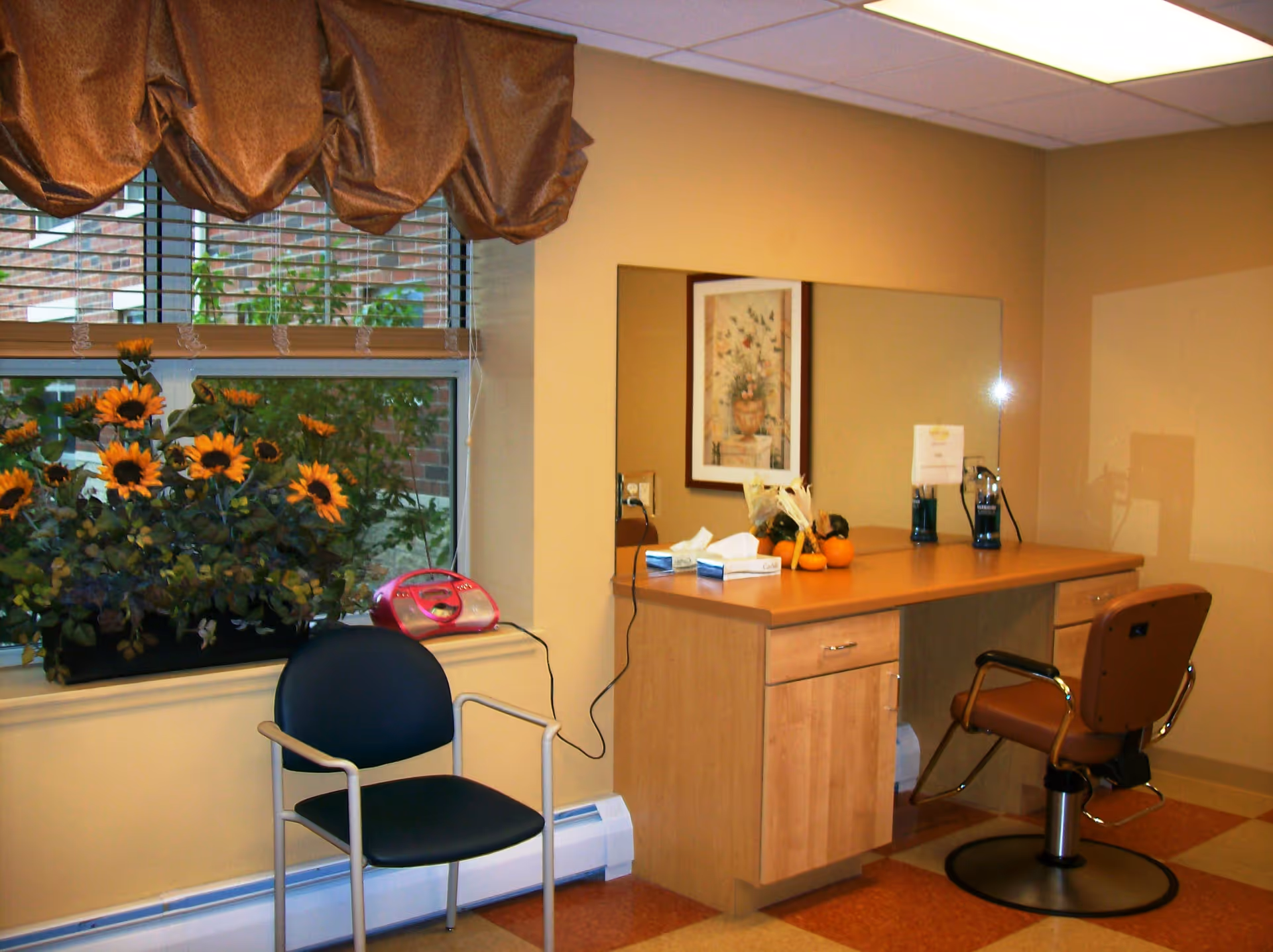 Interior room with a wooden vanity desk and a large mirror on the wall. On the desk are tissue boxes, small decorative pumpkins, and two blue containers. In front of the desk is a brown salon-style chair. To the left, there is a black chair with metal armrests next to a window with blinds and a brown valance. A planter with sunflowers is placed on the windowsill. The floor has a checkered pattern with orange and beige tiles.