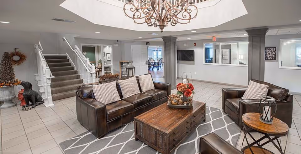 A spacious and well-lit common area with a brown leather sofa and armchairs arranged around a wooden coffee table on a patterned rug. There is a large chandelier hanging from the ceiling, a staircase with white railings, and a TV mounted on the wall. The room has tiled floors and neutral-colored walls with decorative accents.