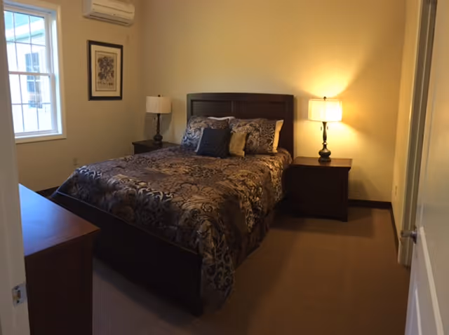 A bedroom with a double bed topped with patterned bedding, two bedside tables with lamps, a dresser, and a window.