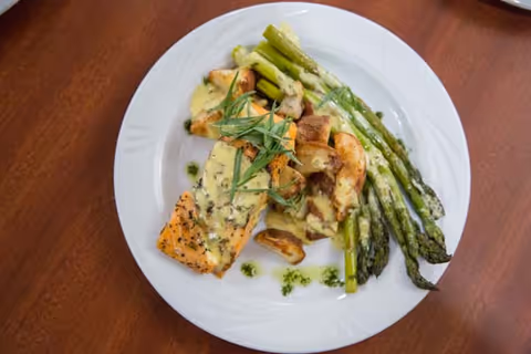 A white plate with a serving of cooked salmon topped with a creamy herb sauce, roasted potatoes, and steamed asparagus on a wooden table.