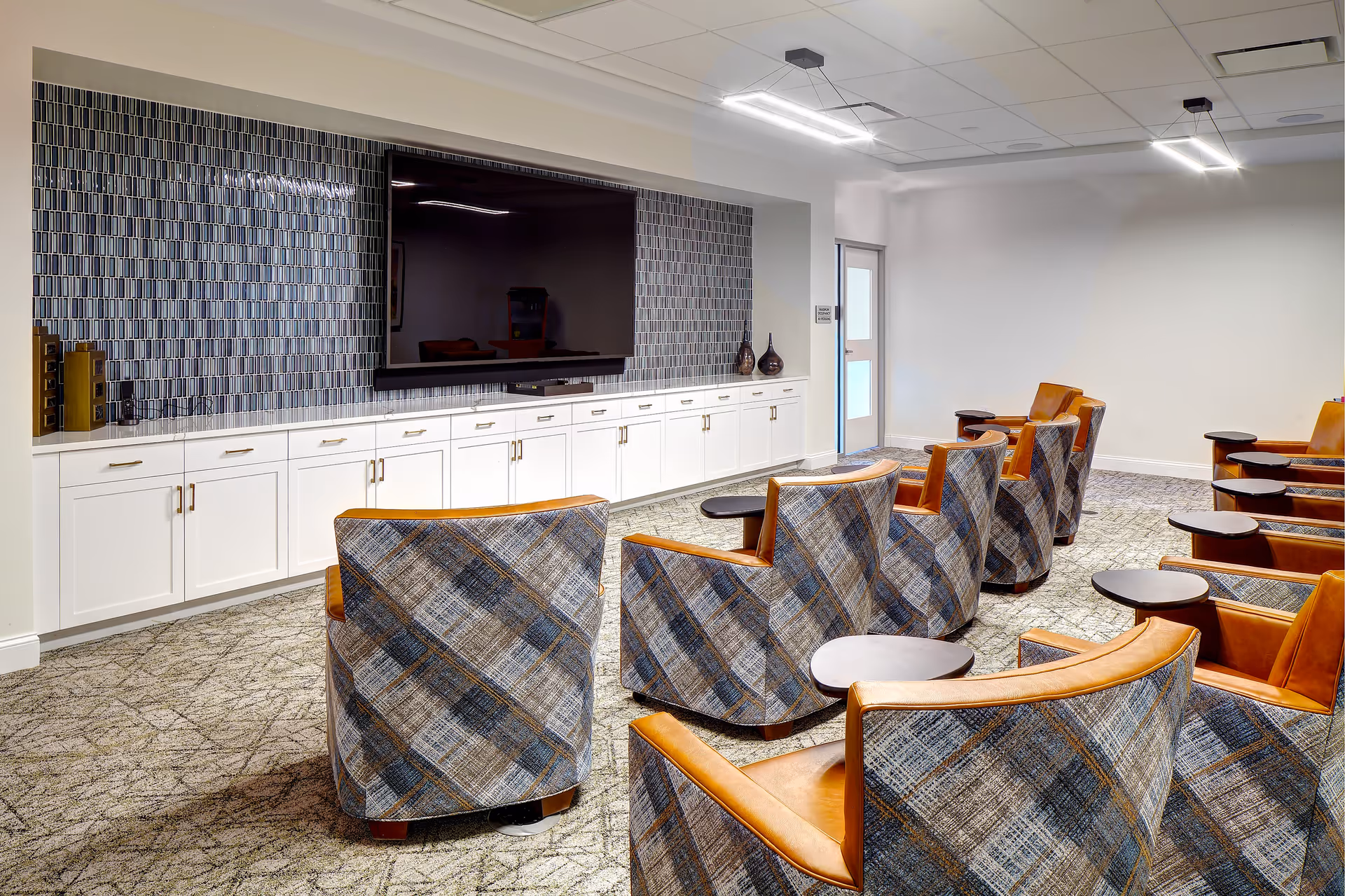 Rows of upholstered chairs with small tables facing a large wall-mounted TV above white cabinetry in a bright common room.