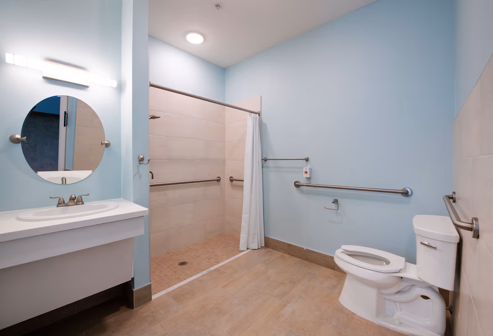 A spacious bathroom with light blue walls featuring a white sink with a round mirror above it, a walk-in tiled shower with grab bars and a white shower curtain, and a white toilet with grab bars on the walls around it.