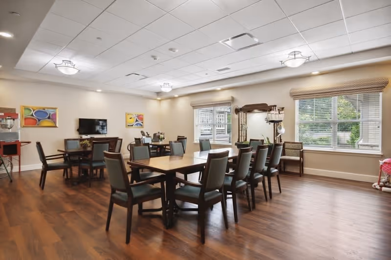 A spacious dining room with multiple wooden tables and chairs arranged neatly. The room has large windows with blinds, allowing natural light to brighten the space. The walls are decorated with colorful abstract paintings, and there is a television mounted on one wall. The floor is wooden, and the ceiling has recessed lighting and decorative light fixtures.