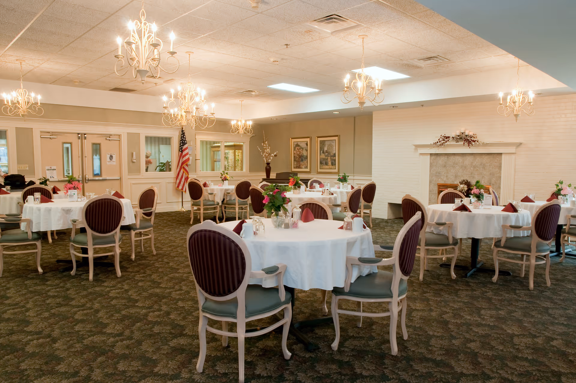 A dining room with round tables covered in white tablecloths, each set with glassware, napkins, and floral centerpieces. The room features multiple chandeliers hanging from the ceiling, an American flag near the back wall, framed artwork, and a decorative fireplace. The chairs have cushioned seats and backs with a striped pattern.