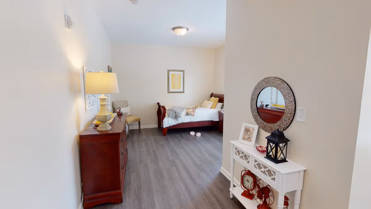 A senior living bedroom with a wooden bed against the far wall, decorated with pillows and a blanket. To the left, there is a wooden dresser with a lamp and decorative items on top. A beige chair is placed next to the dresser. On the right side, a white console table holds a framed picture, a black lantern, and other decorative items, with a round mirror hanging above it. The room has light-colored walls and dark wood flooring.