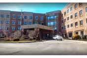 Exterior view of a multi-story brick assisted living facility with a covered entrance, a driveway, and a parked car in front. The building has multiple windows and an American flag near the entrance.