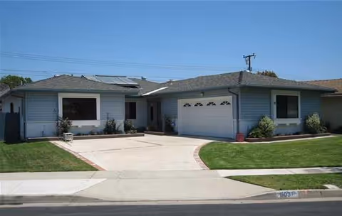 Single-story residential building with a light blue exterior, a two-car garage, and a driveway. The front yard has well-maintained green grass and small shrubs along the house. The sky is clear and blue.