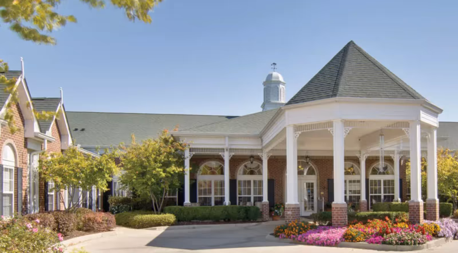 Front entrance of a brick senior living building with a covered porte-cochère, white columns, cupola, and landscaped flowerbeds.