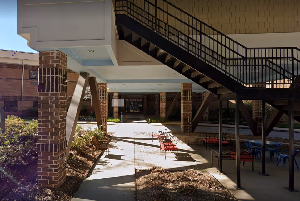 Outdoor covered seating area with benches and chairs with red cushions under a staircase, surrounded by brick pillars and landscaping, leading to a building entrance with a 'Welcome Back' sign.