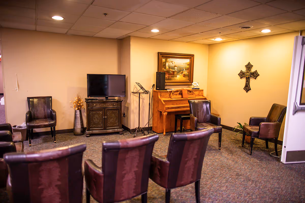 A cozy common area with several brown leather chairs arranged in a semi-circle facing a TV on a wooden cabinet. There is a piano with a framed painting above it and a decorative cross on the wall. The room has beige walls and a carpeted floor.