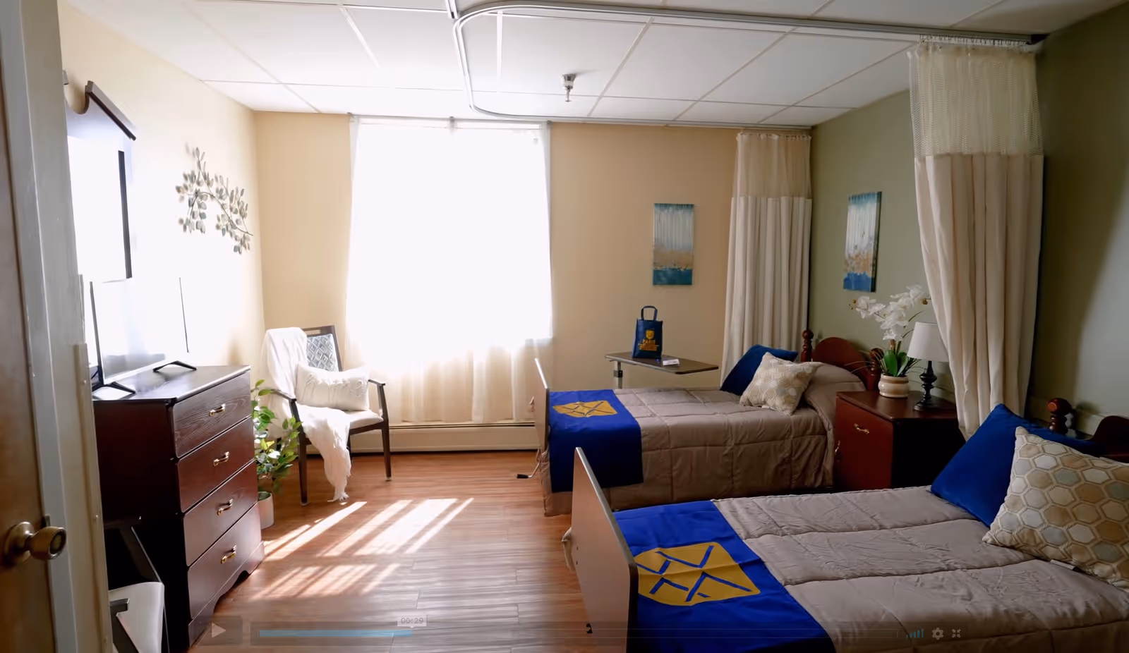 Sunlit shared senior care bedroom with two beds, nightstands, a dresser with TV, and a chair by the window.