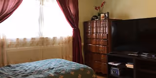 A bedroom with a bed covered in a blue patterned blanket, a window with sheer white curtains and dark red drapes, a tall wooden dresser, and a flat-screen TV on a stand with shelves holding various items.