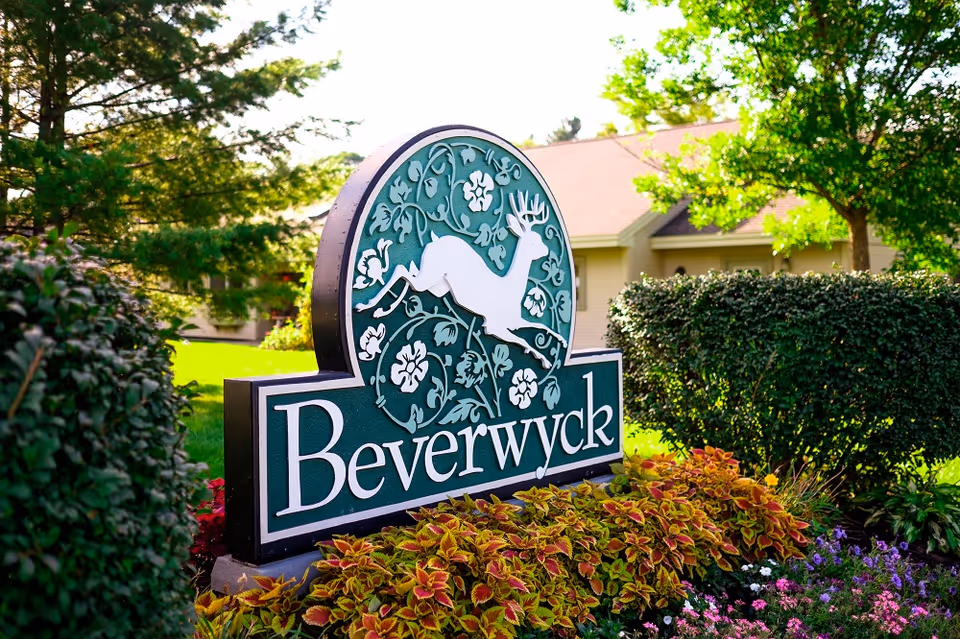 A green and white sign with a leaping deer and floral design that reads 'Beverwyck' surrounded by colorful plants and bushes, with a building and trees in the background under bright sunlight.