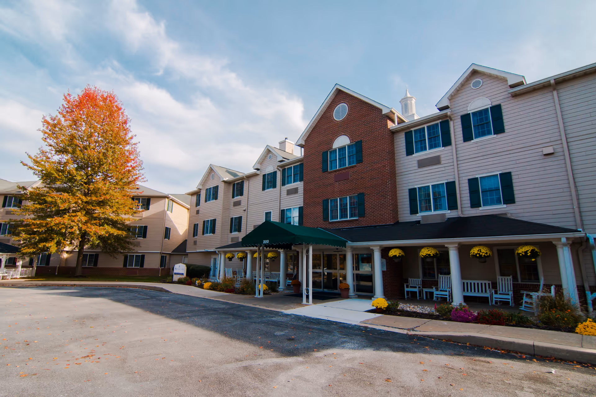 Front exterior of a multi-story senior living building with a covered entrance, porch rocking chairs, and a tree with autumn foliage.