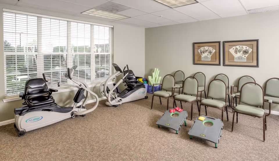 A small exercise room with two NuStep recumbent cross trainers near a large window with white blinds. Several green chairs are arranged along the wall, and two cornhole boards with bean bags are set up on the carpeted floor. Two framed artworks hang on the wall above the chairs.